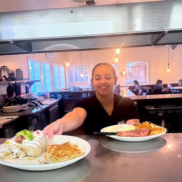 A smiling Waffle Shop server presents two plates of breakfast burritos and hash browns.