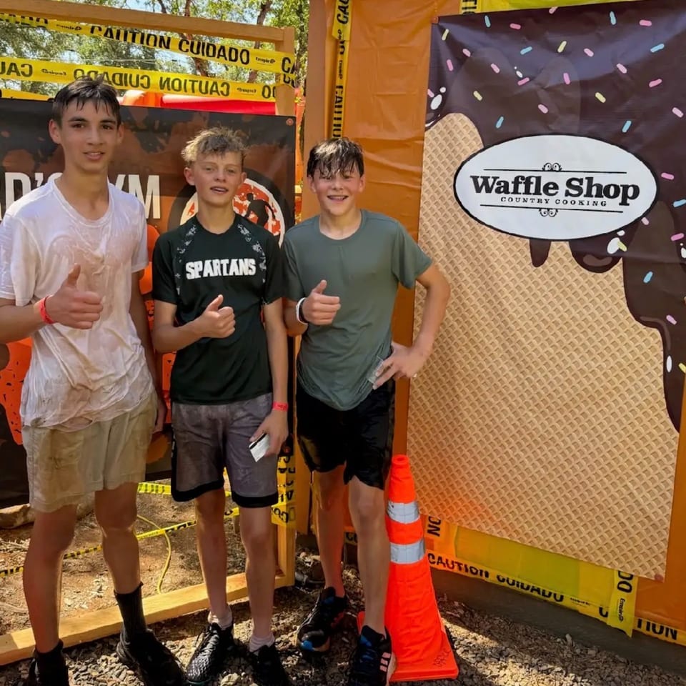 Three muddy boys give thumbs up in front of a Waffle Shop banner at an outdoor event.