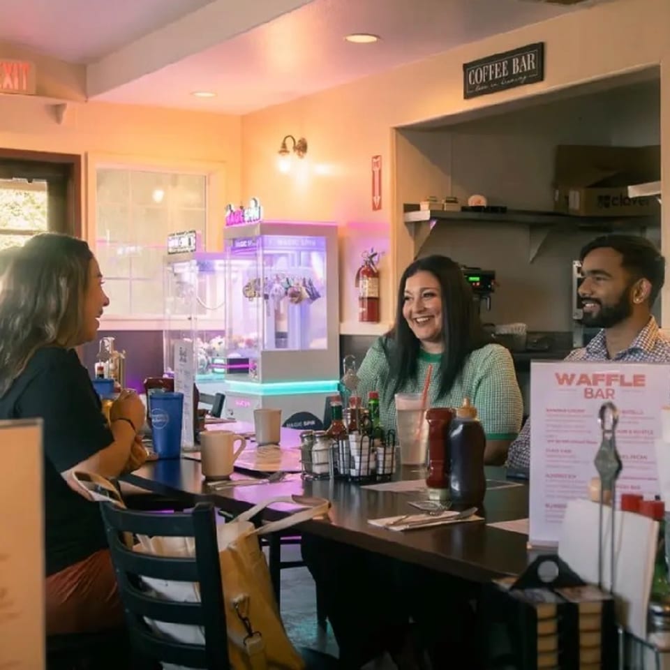 Three people smiling and talking at a diner counter with a "Coffee Bar" sign above.