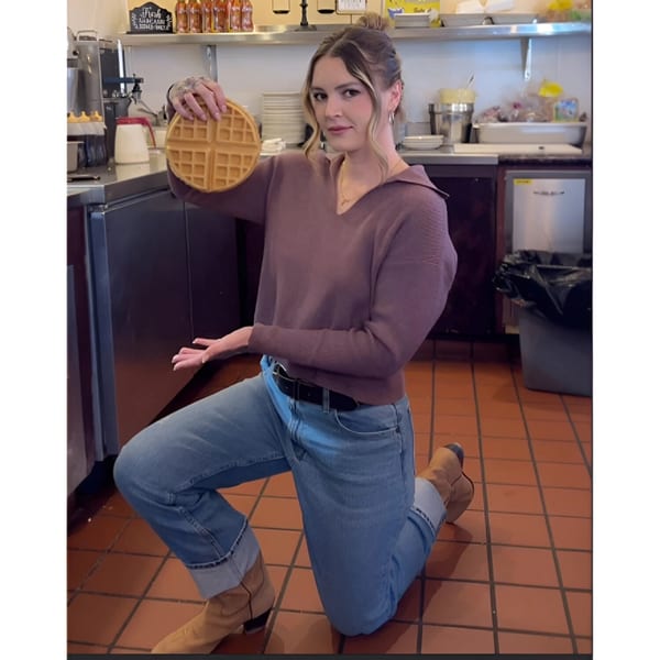 A woman kneels on a tiled floor in a Waffle Shop kitchen, holding up a round waffle.