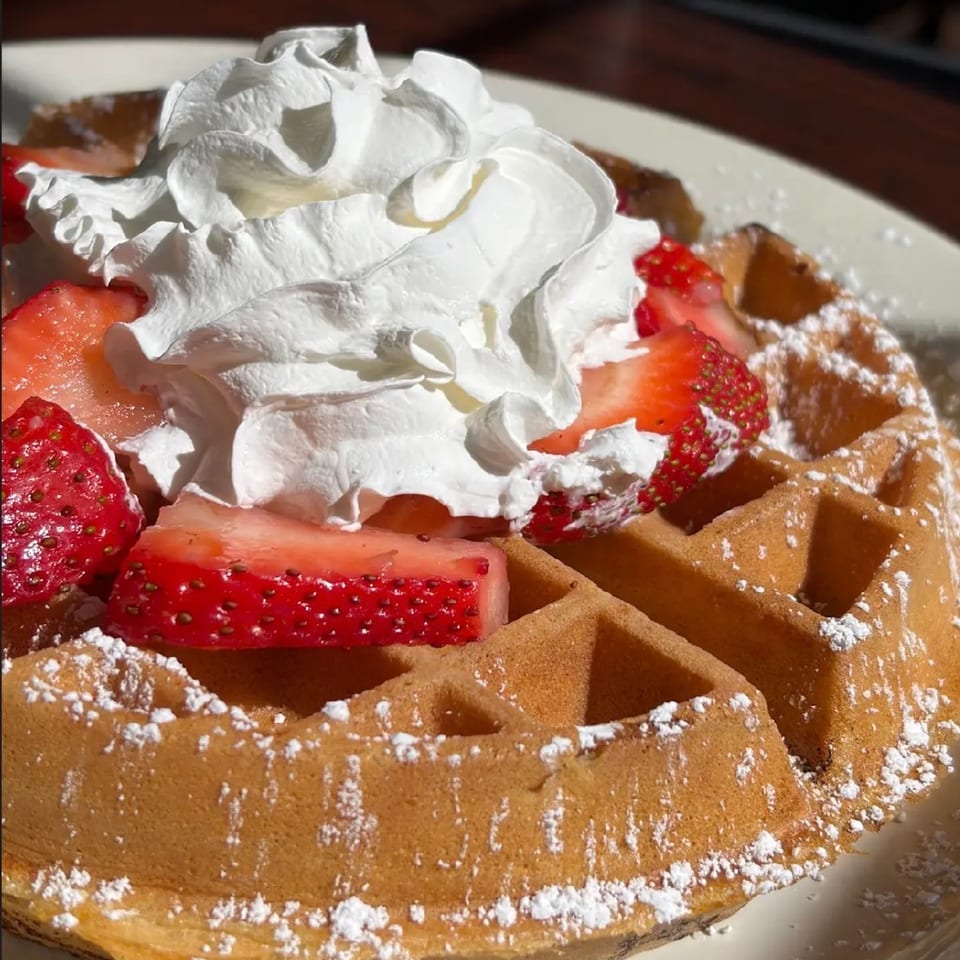 Golden waffle topped with fresh strawberries, whipped cream, and powdered sugar on a white plate.