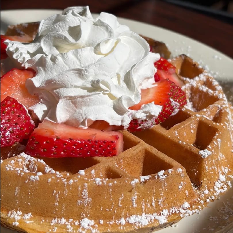 Golden waffle topped with fresh strawberries, whipped cream, and powdered sugar on a white plate.