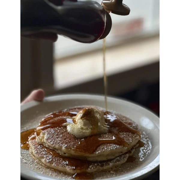 Syrup being poured onto a stack of pancakes with a scoop of butter and powdered sugar.