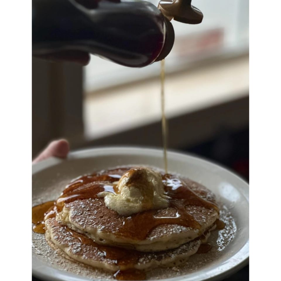 Syrup being poured onto a stack of pancakes with a scoop of butter and powdered sugar.