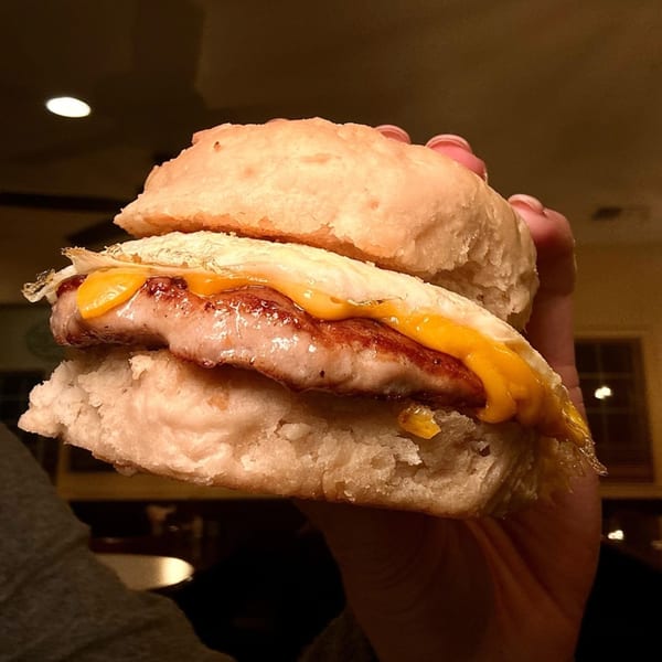 A hand holds a breakfast sandwich with sausage, egg, and melted cheese on a biscuit.