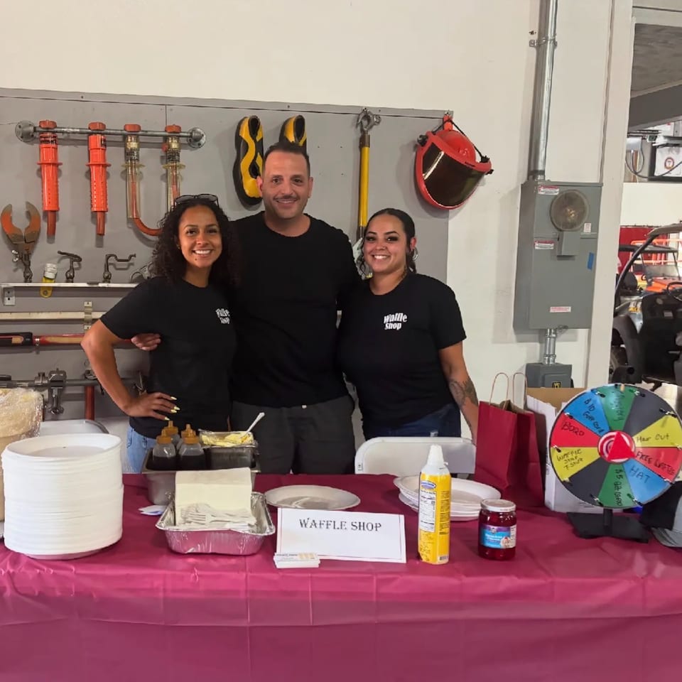 Three smiling people in "Waffle Shop" shirts stand behind a table with waffle toppings and a prize wheel.