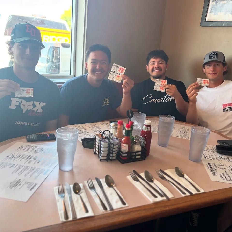 Four young men smiling and holding up raffle tickets at a restaurant table with menus and drinks.