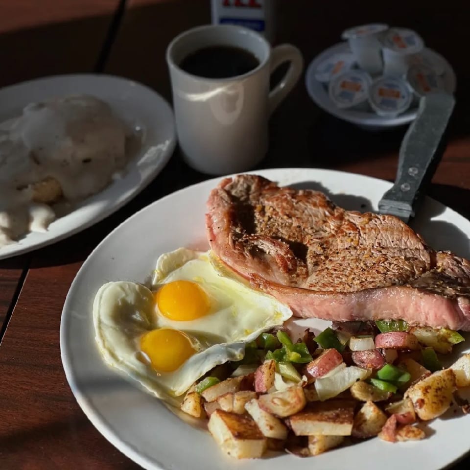 Steak and eggs breakfast with home fries, coffee, and biscuits and gravy on a wooden table.
